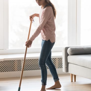 Full-length image of barefoot young woman stands in living room homeowner doing house chores cleaning wooden laminate floor using microfiber wet mop pad, housekeeping job or routine home work concept
